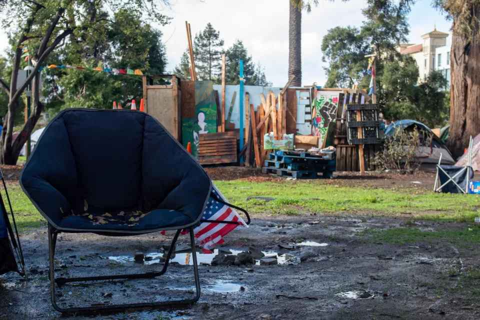 Empty navy blue camping chair with small American flag in front of unhoused encampment in People's Park, Berkeley, California.